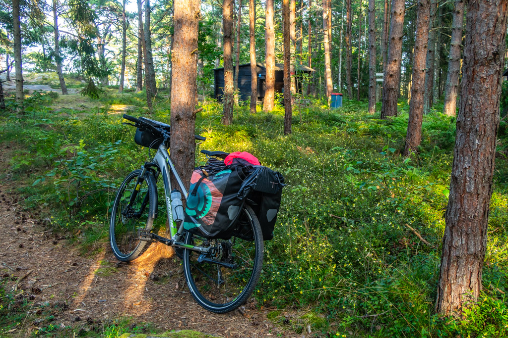 Cykel packad med tält och annat står lutad emot ett träd i fin tallskog på Gränsö utanför Västervik. Ett vindskydd skymtar i bakgrunden.