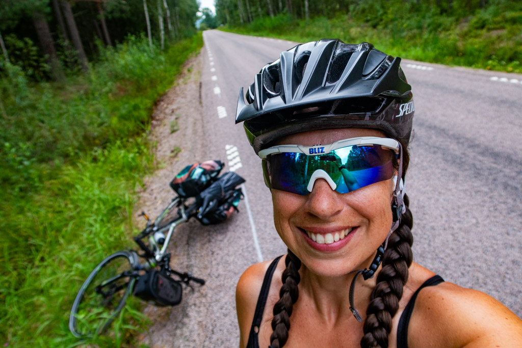 Pernilla Lindblom med hjälm och solglasögon tar selfie vid sidan av väg. Cykel ligger vid sidan om.