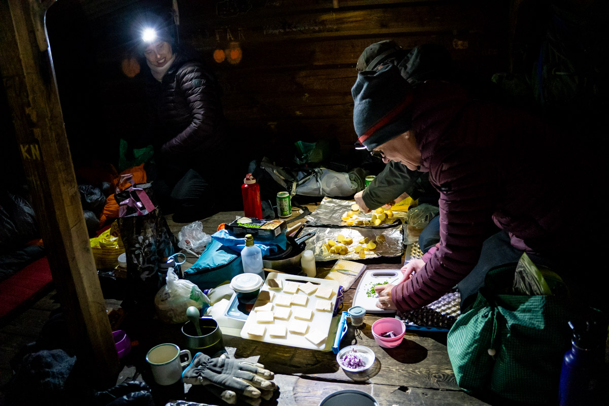 Utesovare förbereder matlagning i vindskydd i mörker, med pannlampor som lyser upp. 